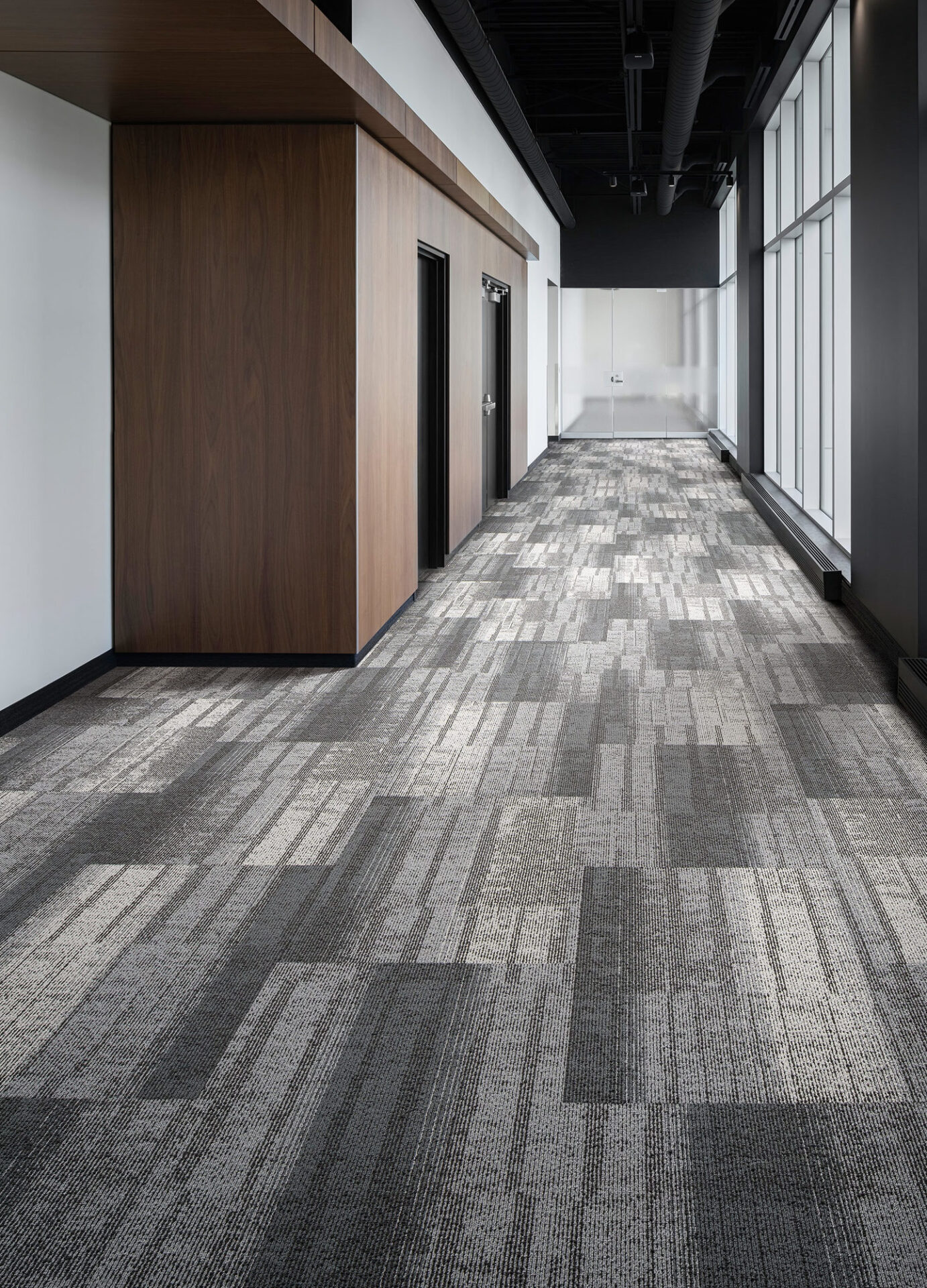 Gray and charcoal linear carpet tile running down a long office corridor beside glass walls