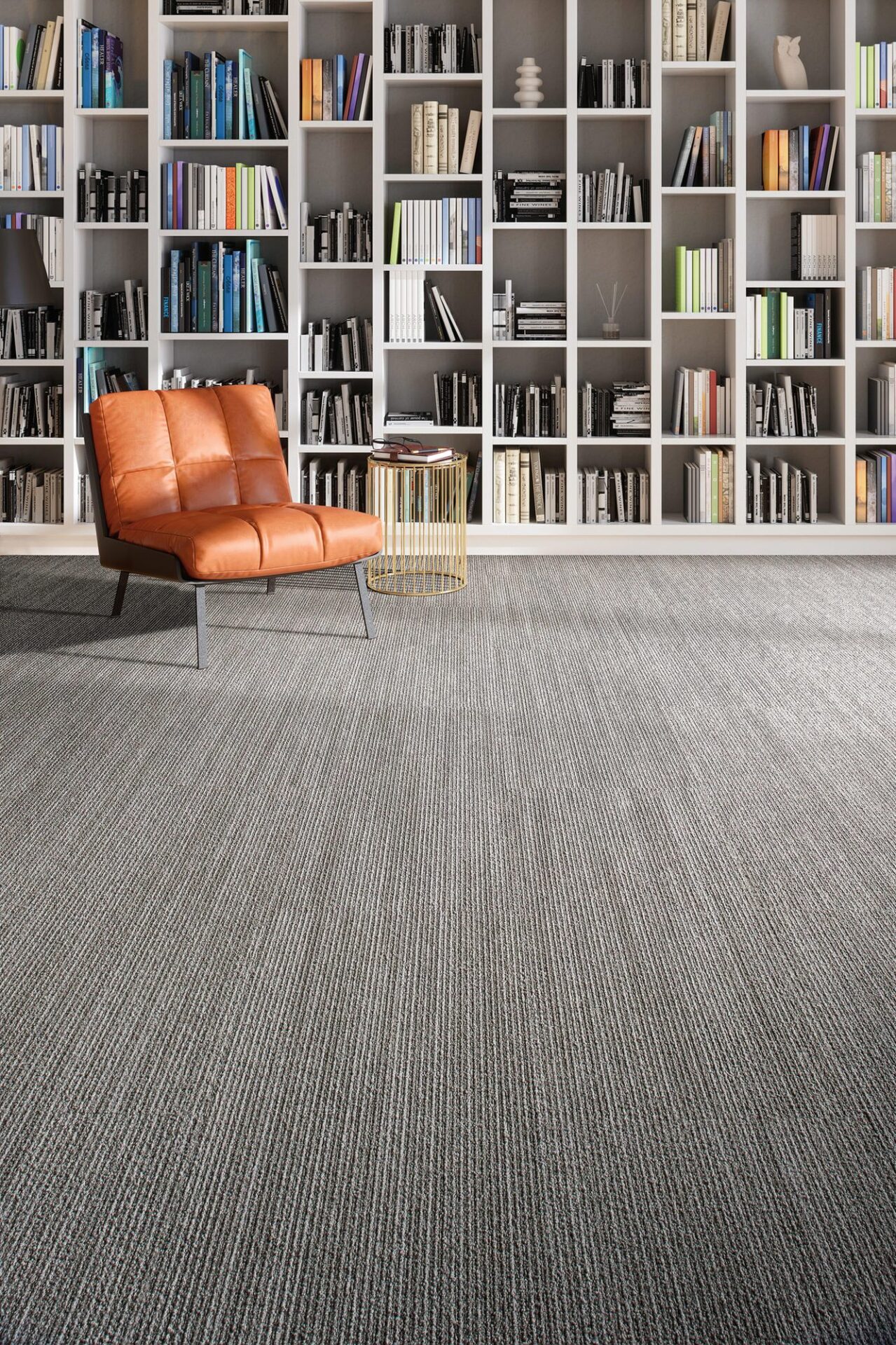 Library reading area with gray linear carpet, an orange armchair and a wall of filled bookshelves