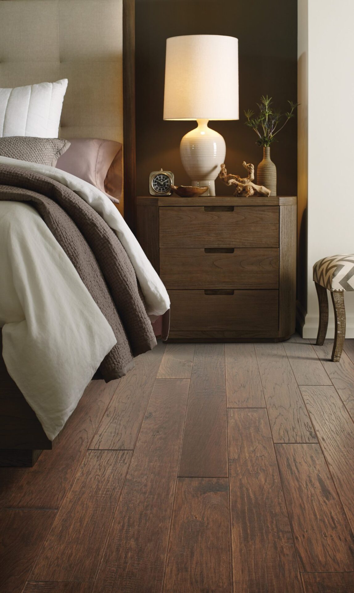 Bedroom close-up of hand-scraped brown hickory hardwood beside a bed and wood nightstand with a white lamp