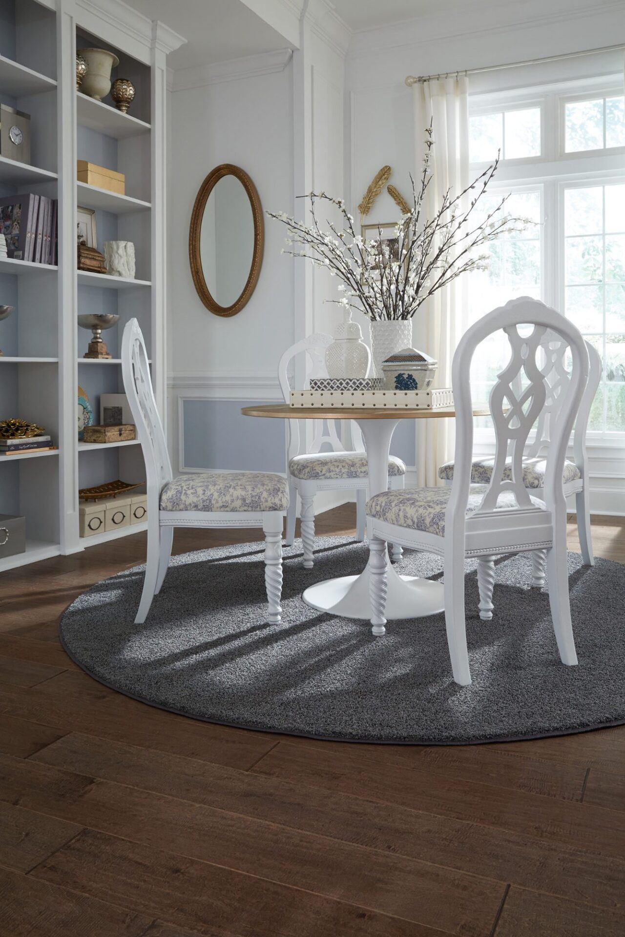 Dining area with warm brown maple hardwood, a round white table and chairs on a gray rug beside built-in shelves