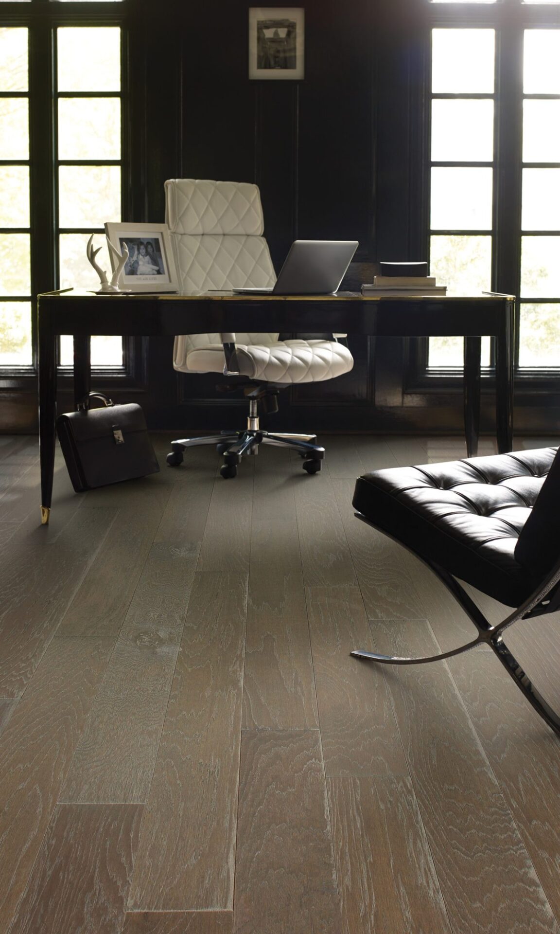 Home office with gray-brown hickory hardwood, a dark wood desk, white desk chair and black leather lounge chair