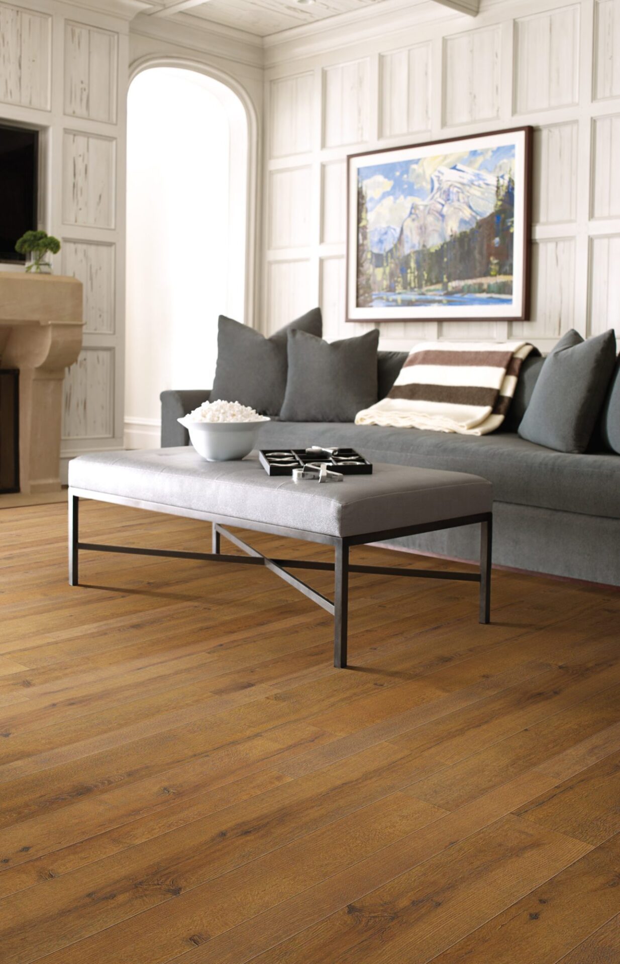 Living room with warm brown wood-look laminate, a gray sofa, upholstered ottoman and white paneled walls