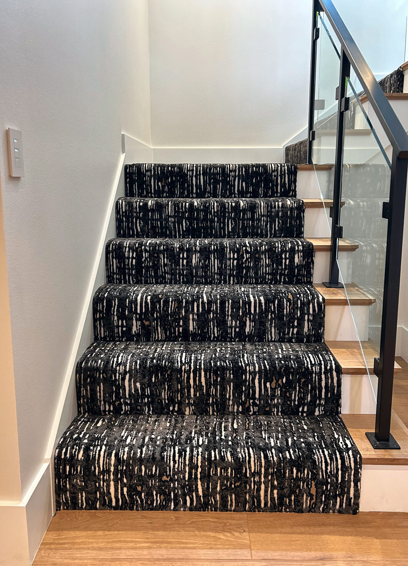 Patterned black and white carpet on stair treads beside a glass railing and light oak floor