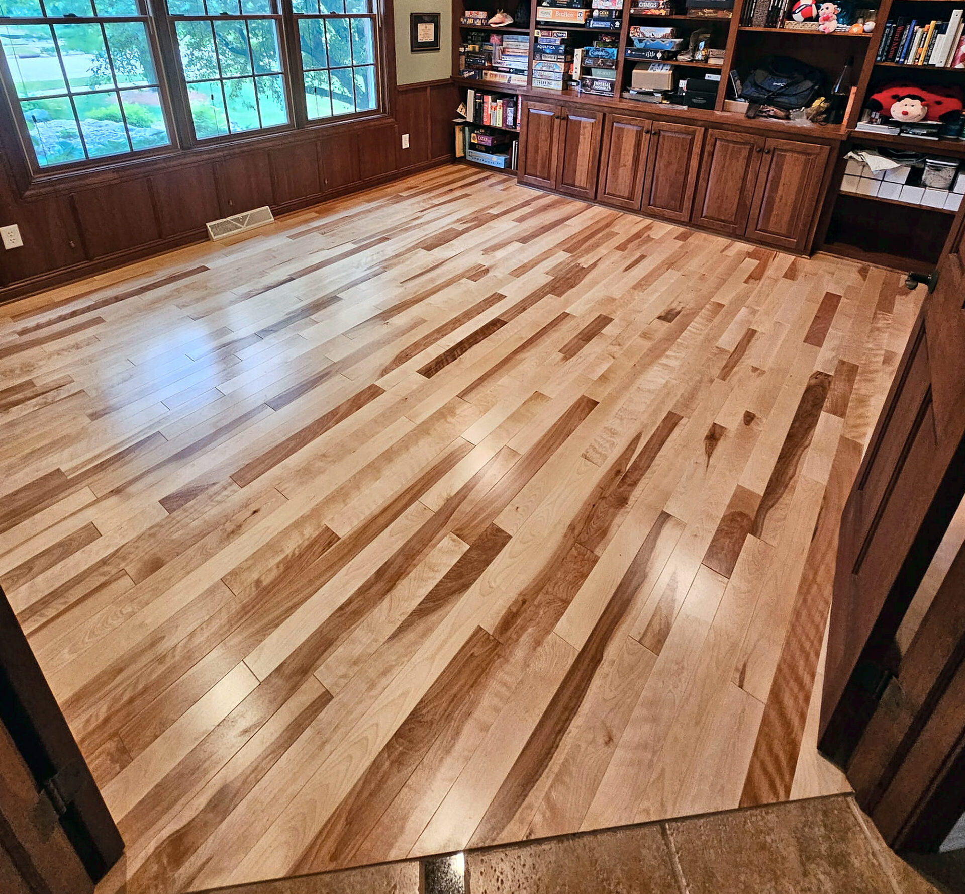 Natural mixed-tone hardwood floor in a den with built-in cabinets and large windows